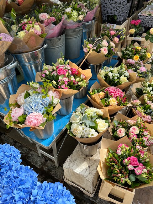 An assortment of fresh floral bouquets arranged in metal buckets and wrapped in brown craft paper, featuring a variety of flowers including pink roses, white hydrangeas, and light green foliage. The b