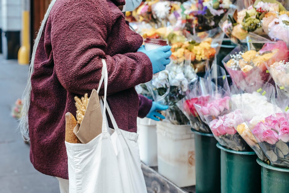 A person wearing a maroon fleece jacket and blue gloves stands beside a display of flower bouquets at Stepney Market. They hold a takeaway coffee cup in one hand and a white tote bag with a baguette a