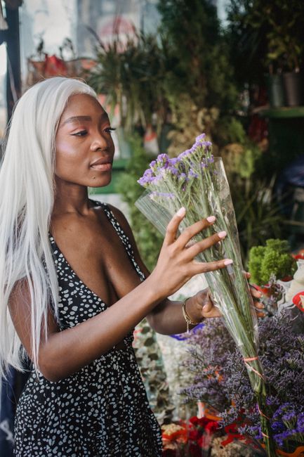 A young woman with long, platinum blonde hair and medium-dark skin holds a tall, clear plastic-wrapped bouquet of purple flowers, likely statice or lavender, at a floral stall. She is wearing a sleeve