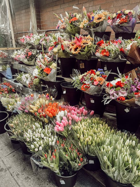 A display of various fresh flower bouquets arranged in black buckets at a flower shop, featuring a variety of colorful blooms including pink and white tulips, orange roses, and pink lilies. The bouque