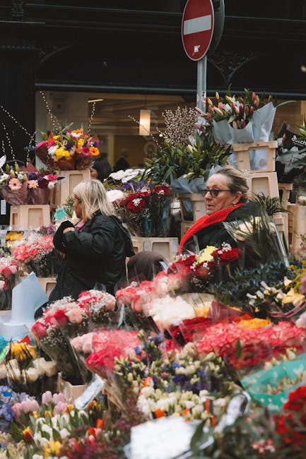 A busy flower market scene featuring a variety of freshly arranged floral bouquets and individual stems wrapped in colorful paper and cellophane. Prominent in the foreground are vibrant pink, red, and